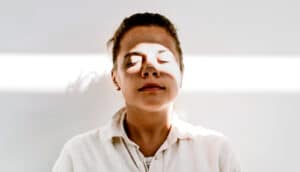 A woman meditates against a white wall with a beam of light shining on her face.