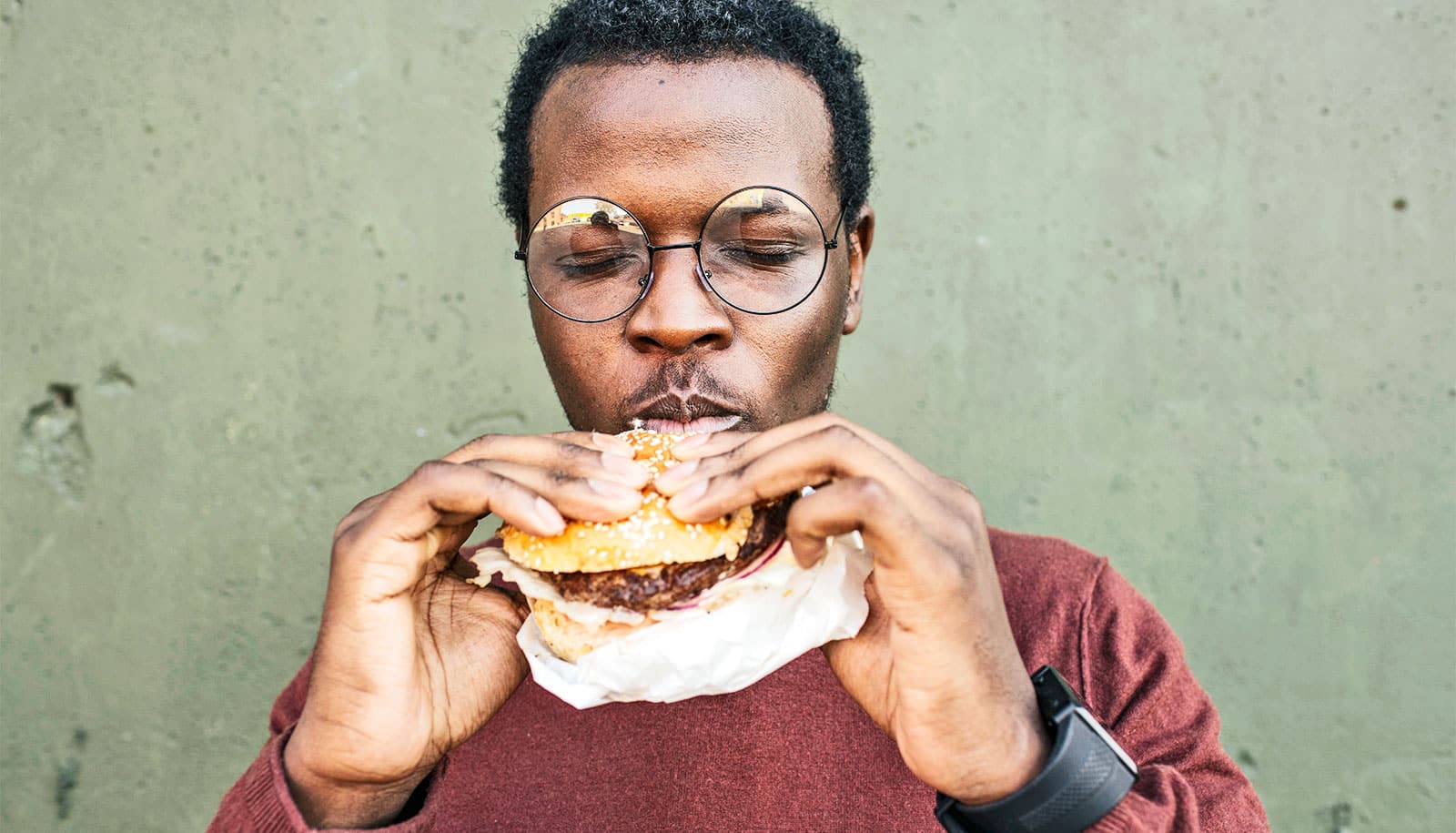 A man wearing glasses closes his eyes while eating a burger outside.