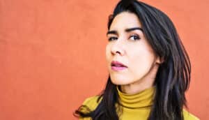 A woman making a complicated facial expression against an orange wall.