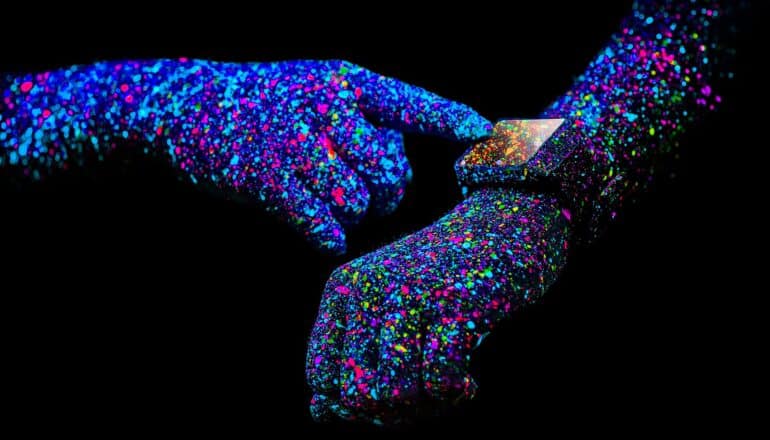 A person uses a smartwatch with their arms covered in specks of colorful paint against a black background.