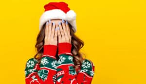 A woman wearing a holiday sweater and Santa hat puts her hands over her face.