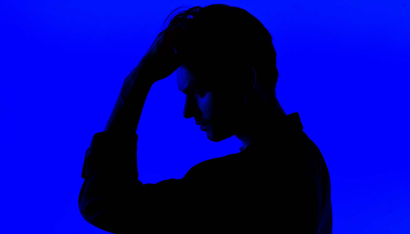 A young man in profile and silhouette holds his forehead.