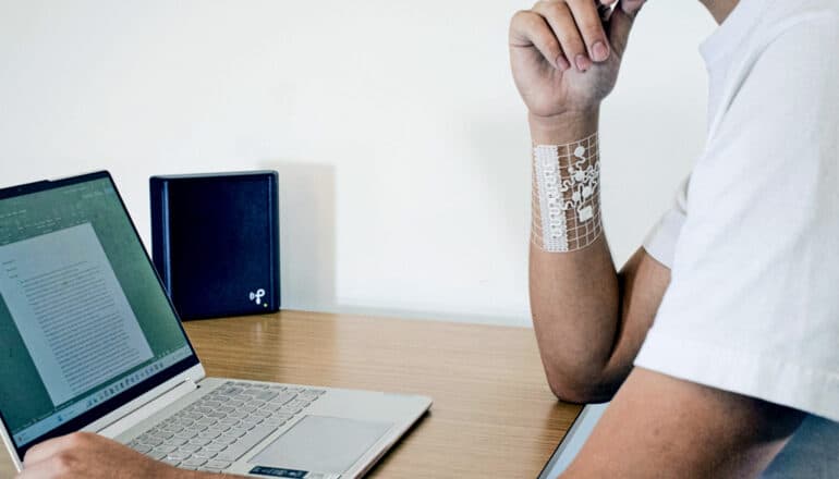 A person working on a laptop at a desk wears the device on their right wrist. It's white and has several wires over a clear patch.