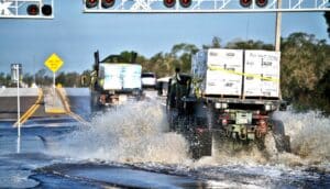 Trucks carrying pallets of water and rations drive through splashing floodwater on a highway.
