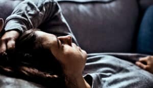 A woman lays on a couch with her hand on her head.