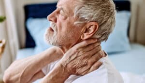 A man sitting on his bed grasps his neck with both hands and winces in pain.