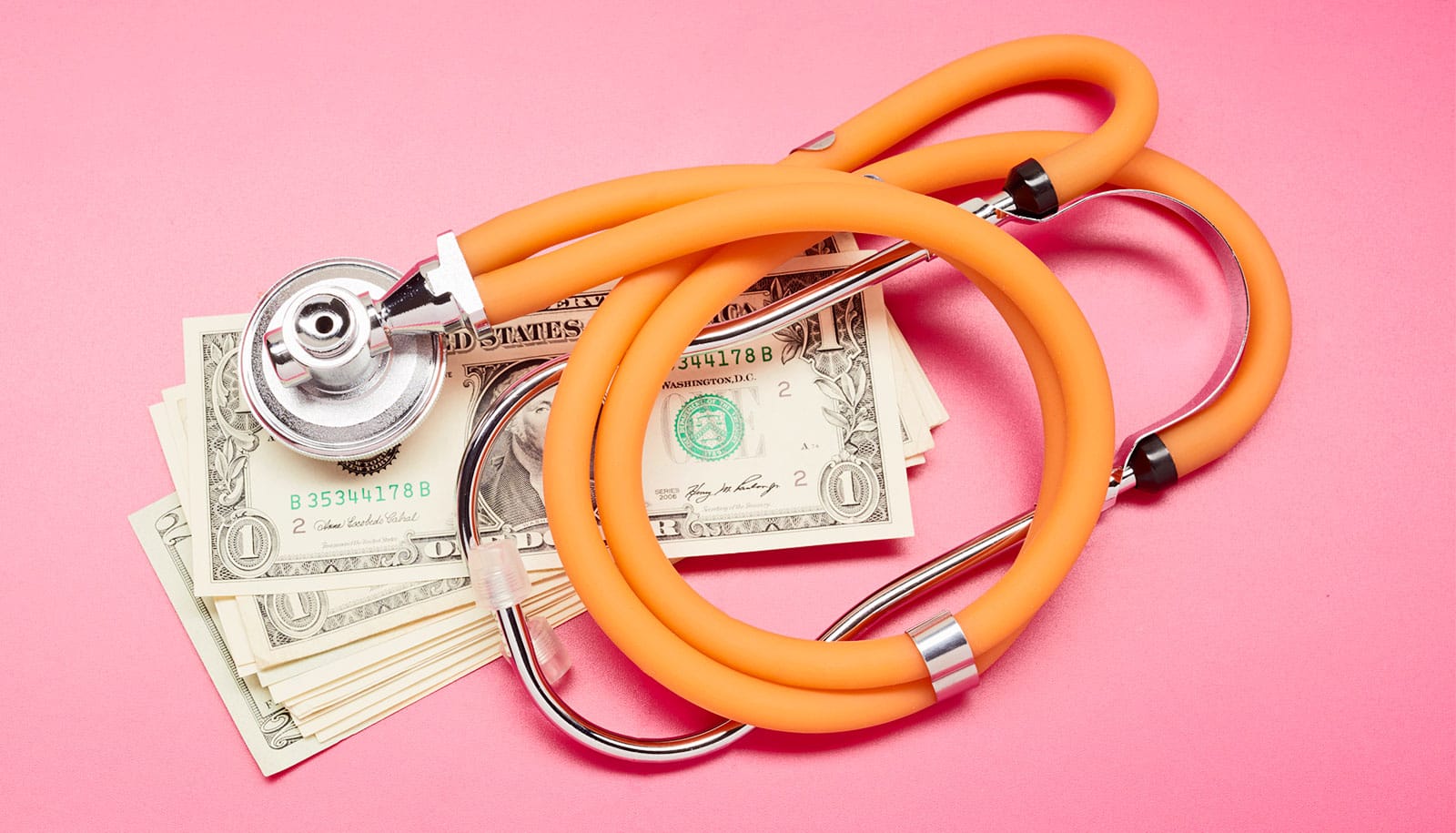 An orange stethoscope over a stack of dollar bills on a pink background.