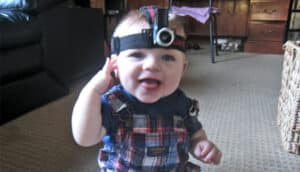 A baby wears a head-mounted camera while sitting on the floor and smiling.