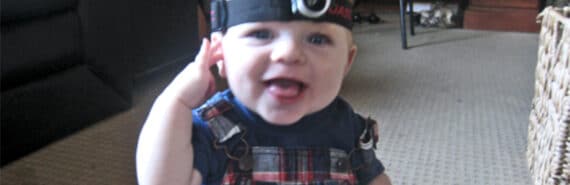 A baby wears a head-mounted camera while sitting on the floor and smiling.