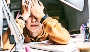 A high school student sitting at a desk doing homework puts her hands over her face.