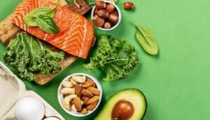 Foods that make up a keto diet, including avocado, salmon, nuts, eggs, and greens, on a green background.