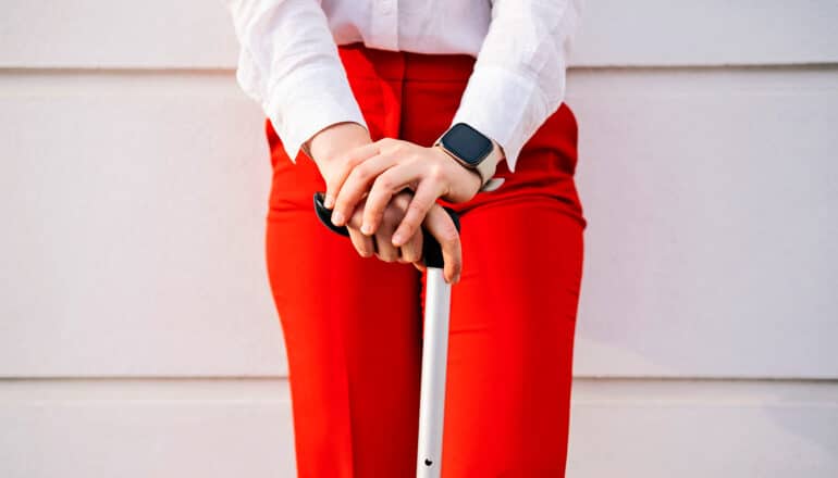 A woman wearing red pants and a smartwatch holds a cane in front of her with both hands.