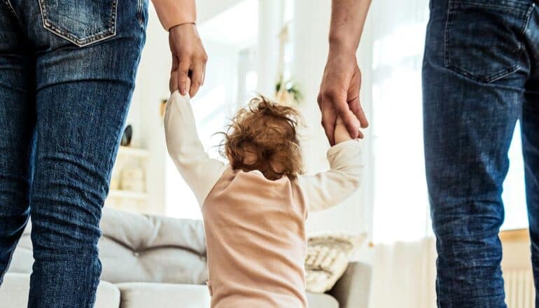Two parents hold their child's hands while at home.