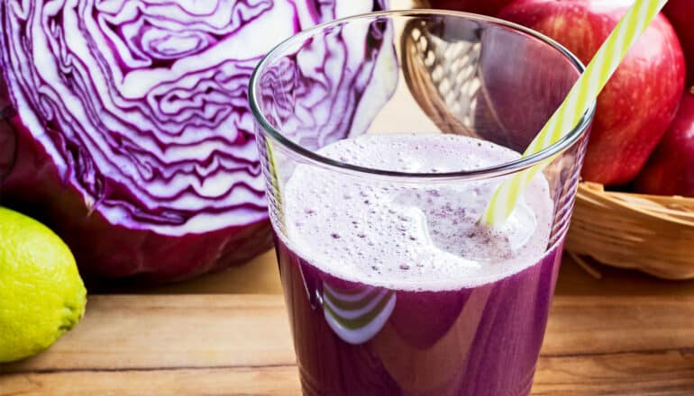 A glass of red cabbage juice sits on a counter next to a red cabbage and a bowl of apples.