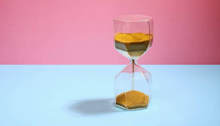 An hourglass stands in front of a blue and pink background.