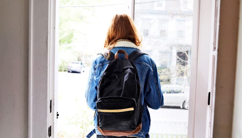 A young woman walks out a front door while wearing a backpack.
