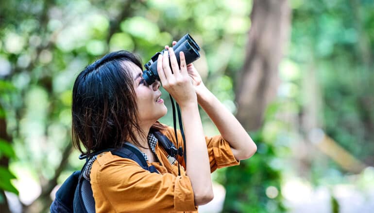 A young woman out in a forest uses binoculars for birdwatching.