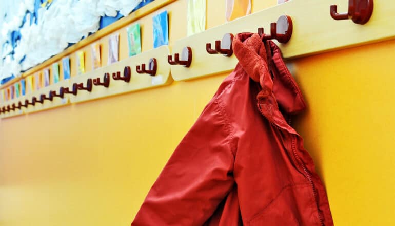 A child's red jacket hangs from a hook in a classroom.