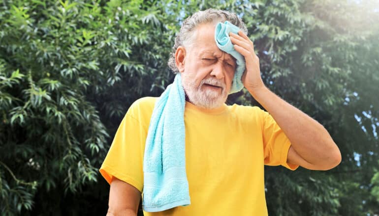 An older man wipes his forehead with a towel as he stands outside.