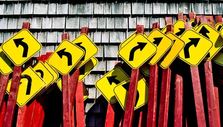 Many black and yellow road signs with arrows pointing in different directions lean against a wooden wall.