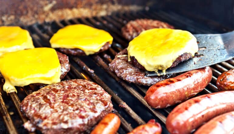 Burgers and hotdogs cook on a grill as the cook removes one burger with cheese on it with a spatula.