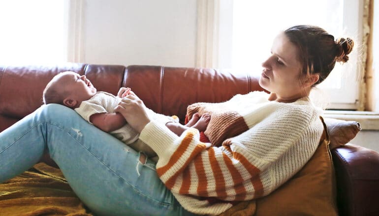 A mother lays on the couch with her baby on her knees while looking depressed.