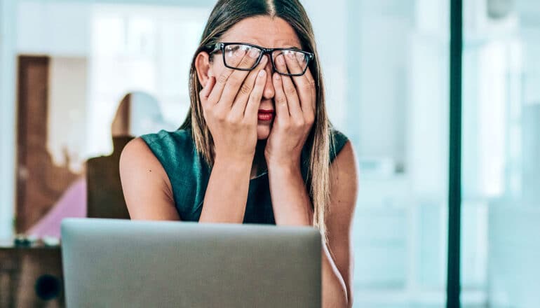 A woman at work reaches under her glasses to put her hands over her face.