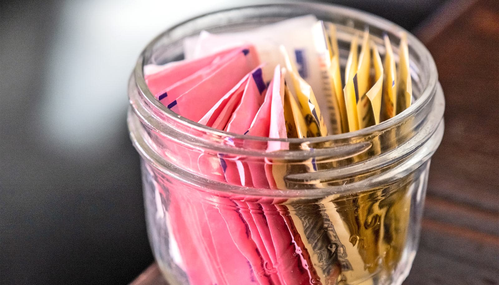 A small jar on a wooden table contains many different packets of artificial sweetener.