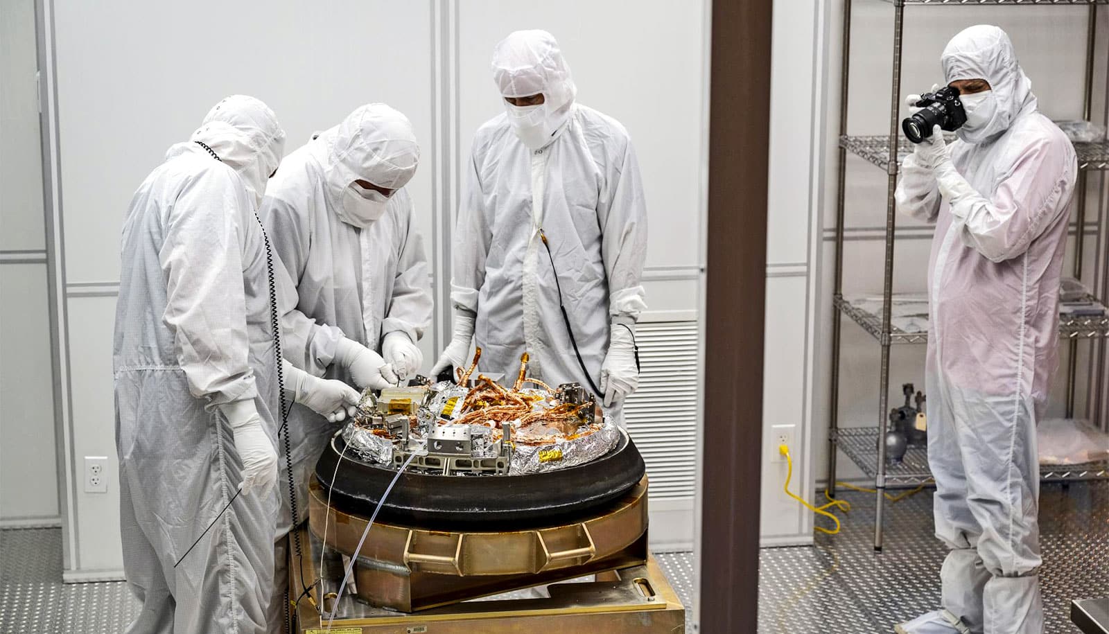 Four scientists in white protective body suits stand over the capsule containing samples from the asteroid Bennu.