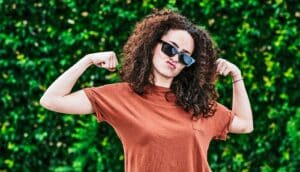 A young woman flexes her biceps and makes a funny face in front of a wall of ivy.