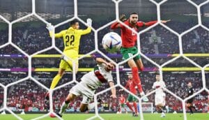 A player heads the ball past a defender and a goalie toward an open net.