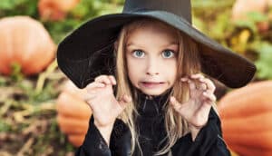 A young girl wears a black witch's costume and makes a scary face while standing in a pumpkin patch.