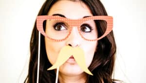 A young woman holds a pair of glasses and fake mustache over her face.