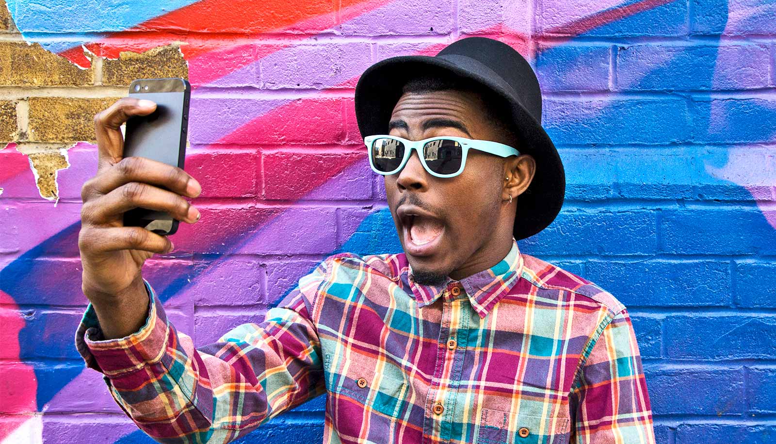 A young man in a colorful plaid shirt, glasses, and a hat takes a selfie in front of a brightly colored wall.