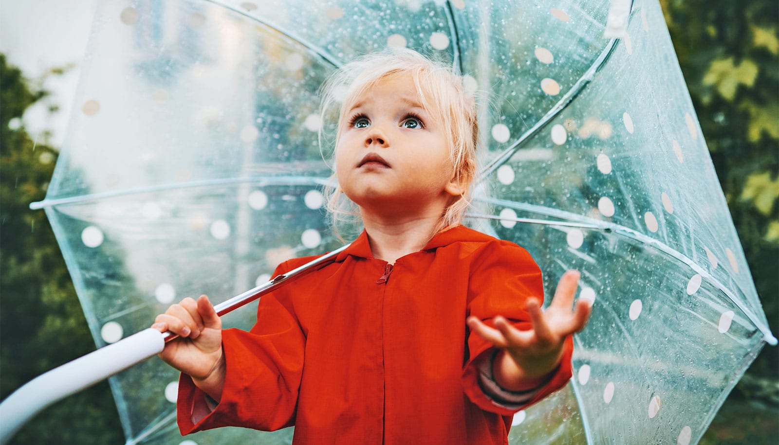 A young girl in a red rain coat holds an umbrella, looks upward, and holds out her hand to check for rain.