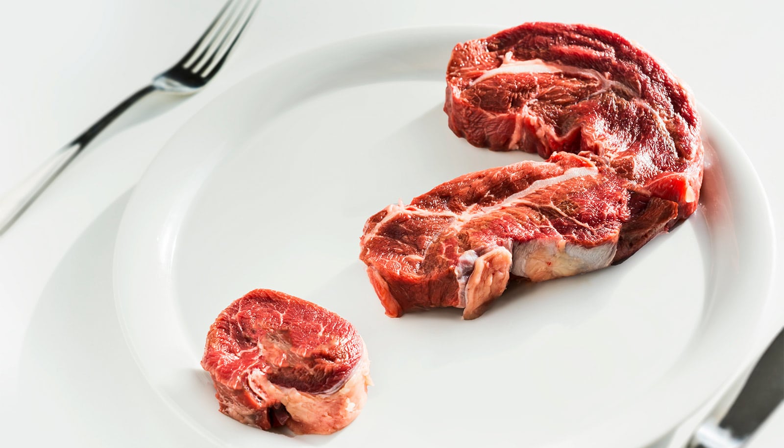 Raw meat in the shape of a question mark sits on a white plate with a fork and knife next to it.
