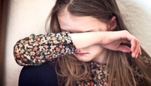 A young girl holds her arm over her face as she sits against a wall.