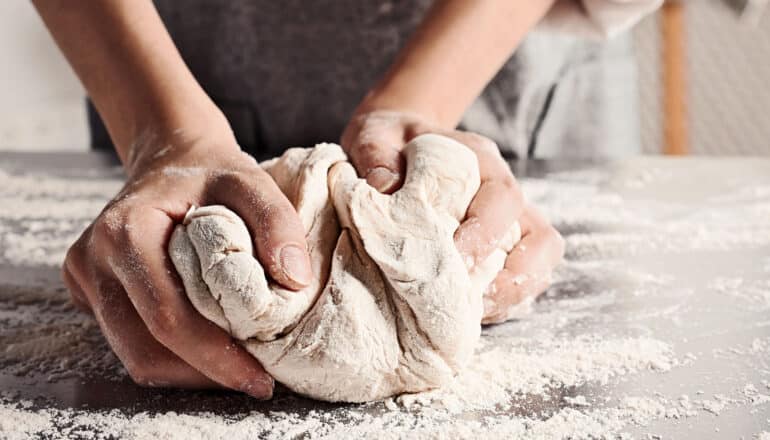 A baker kneads a ball of dough on a metal counter covered in flour.