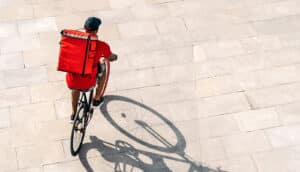 A bike courier rides over cement while carrying a large red package on his back.