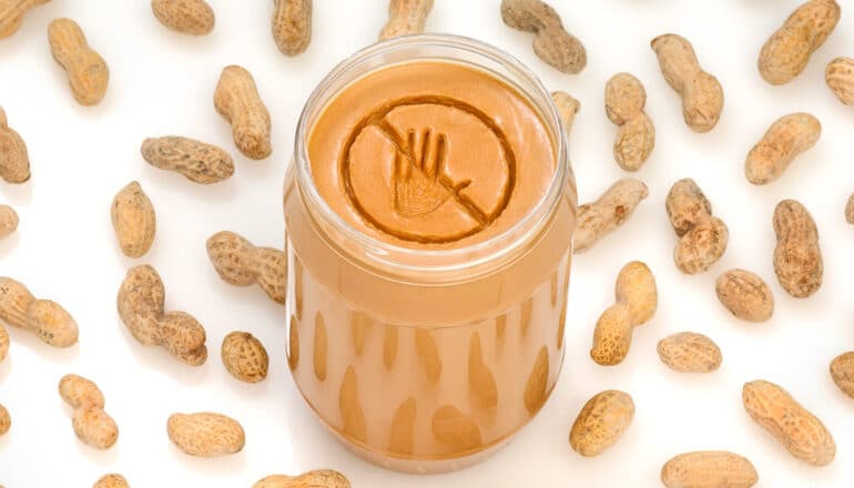 Peanuts on a white surface surround a peanut butter jar with a warning label carved into the peanut butter's surface.