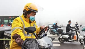 A man wearing a face mask on a motorbike checks his phone while stopped, while white smog fills the air around him.