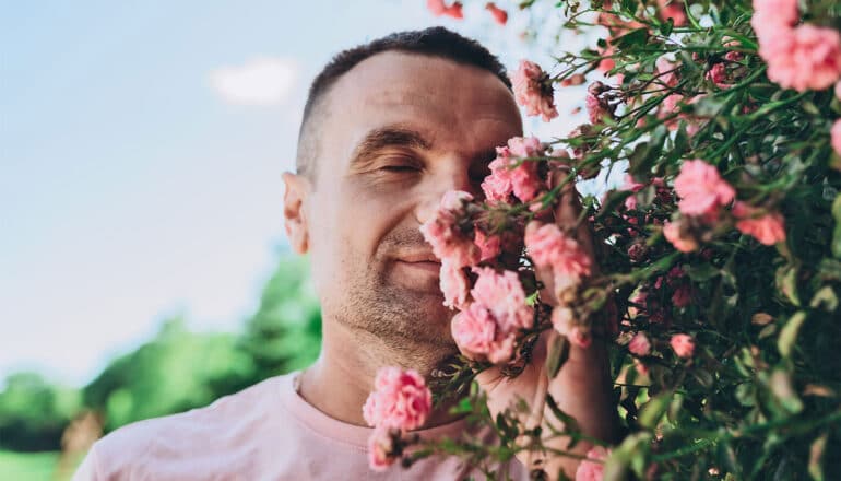 A man closes his eyes and smells pink flowers while standing outside.