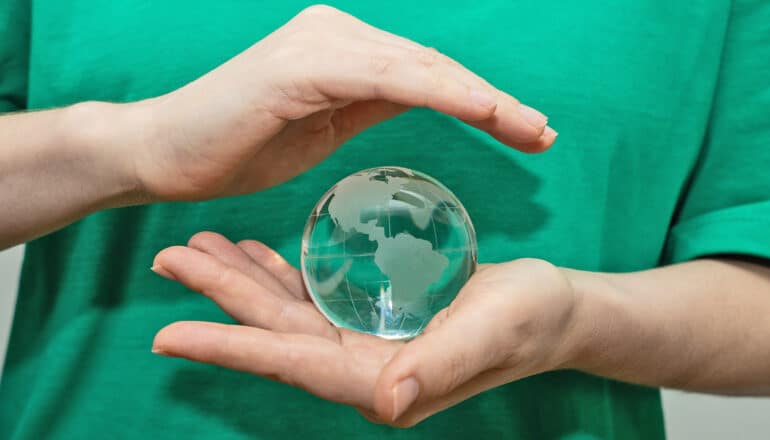 A person holds a glass model of Earth in one palm and covers it protectively with the other hand.