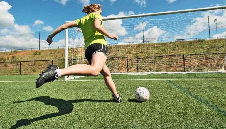 A teen girl runs to kick a soccer ball into an open goal.