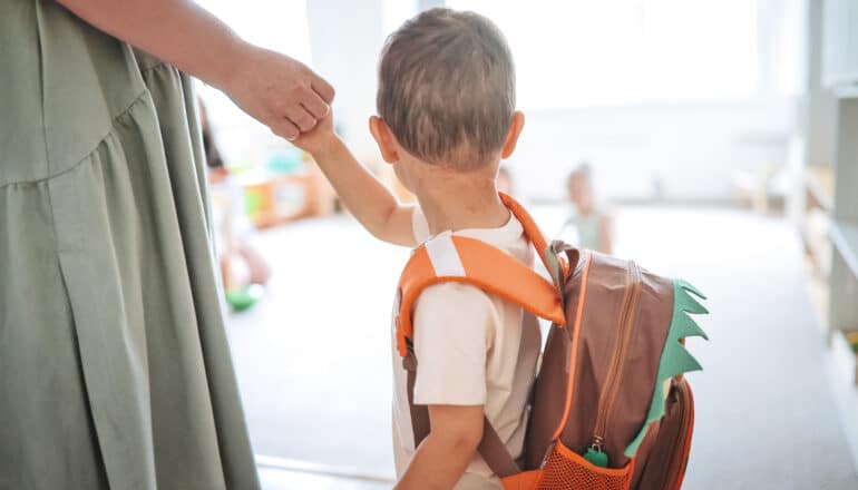 A parent drops a young child wearing a backpack at kindergarten.
