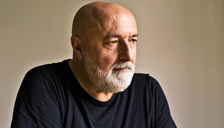 A middle-aged man with a white beard looks off into the middle distance.