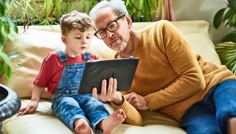 A young boy sits on a couch next to his grandfather holding up a tablet computer.