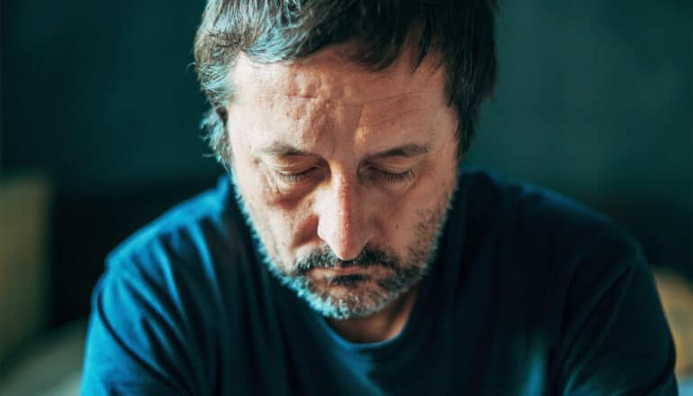 A middle-aged man looks downward while sitting alone.