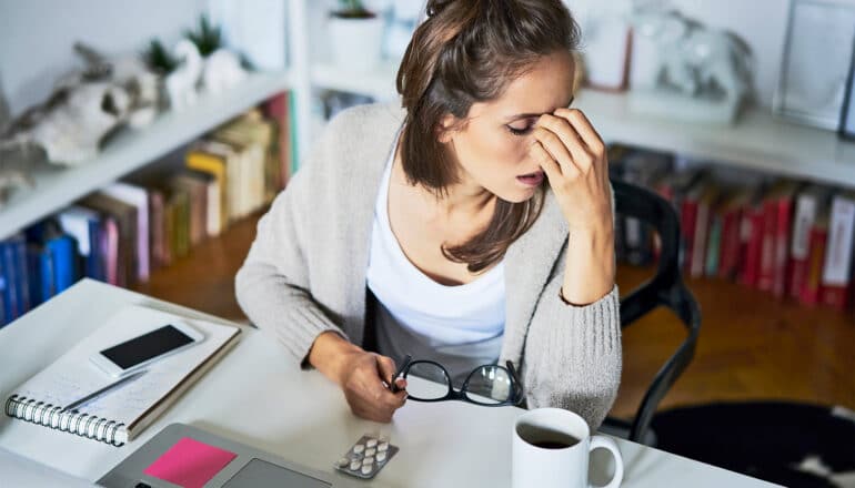 A woman in an office takes off her glasses and grasps the bridge of her nose in pain.