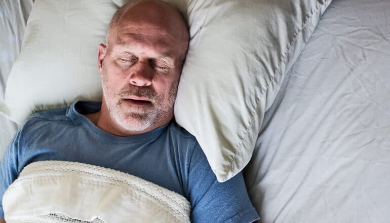A man sleeps with his mouth open while laying on his back in a bed with white sheets.
